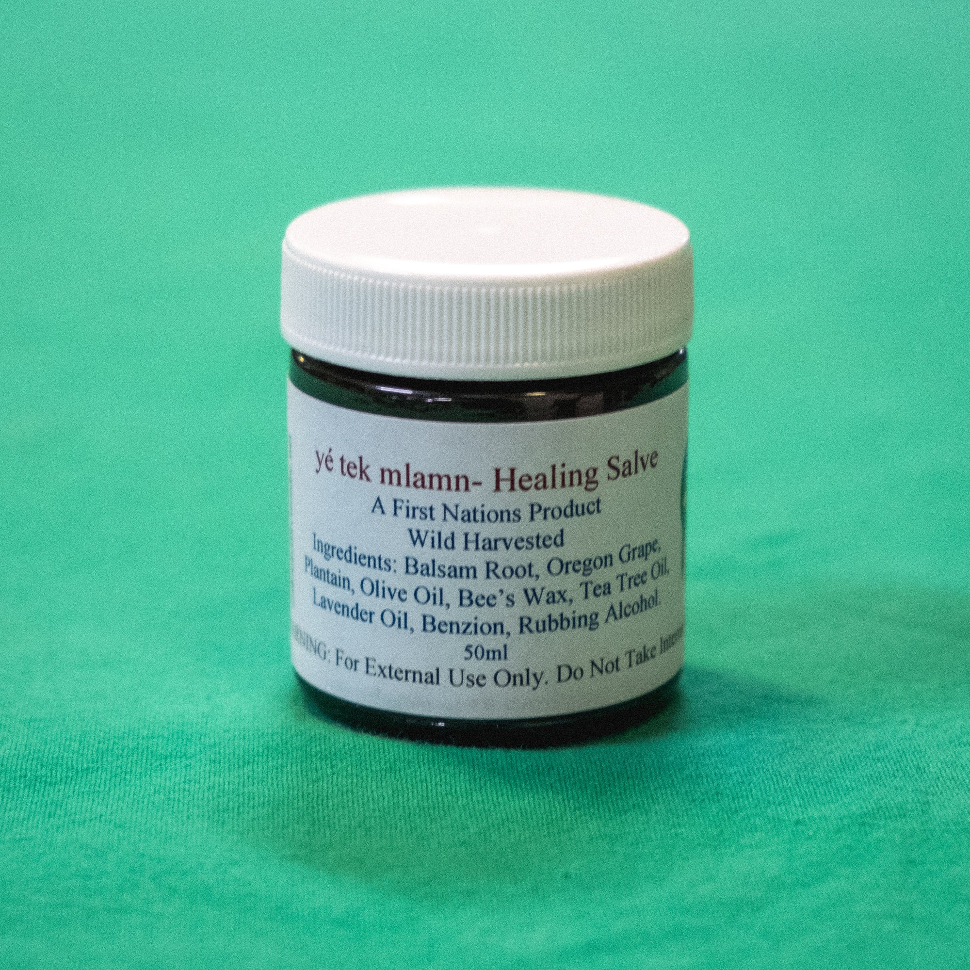 A jar with a label that says "yé tek mlamn - Healing Salve. A First Nations Product. Wild Harvested. Ingrediants: Balsam Root, Organ Grape, Plantain, Olive Oil, Bee's Wax, Tea Tree Oil, Lavendar Oil, Bezion, Rubbing Alcohol. 50ml. Warning For External Use Only. Do not take internally." End of image description.